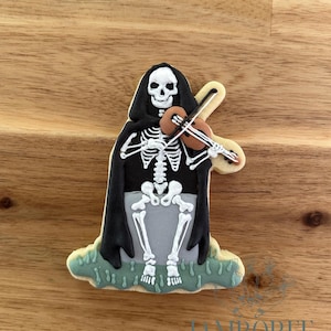 Pode incluir: Um biscoito de Halloween em forma de esqueleto tocando violino. O esqueleto usa uma capa preta e está sentado em uma lápide cinza. O biscoito é decorado com glacê branco, preto, marrom e verde.