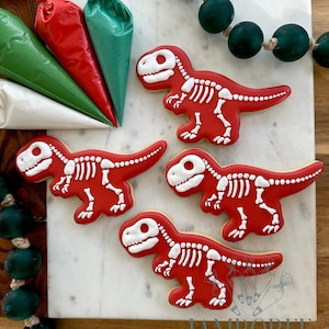 Peut inclure: Quatre biscuits rouges en forme de dinosaure avec des détails d'os blancs sont disposés sur une surface en marbre. Des poches à douille vertes, rouges et blanches sont dans le coin supérieur gauche. Une guirlande de perles vertes est en arrière-plan.