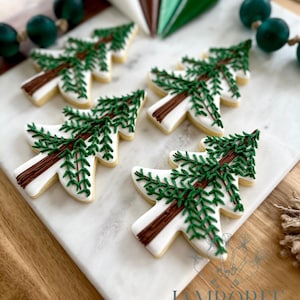 May include: Christmas tree-shaped cookies with white icing, brown trunks, and green pine needles are arranged on a white marble surface. Green beaded garland and piping bags are in the background. The cookies are from Jamboree Home Bakery.