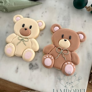 May include: Two bear-shaped sugar cookies decorated with royal icing. One bear is light cream and the other is medium brown. Both feature pink inner ears and paw pads, black eyes and noses, and a sage green piped bow around the neck area.