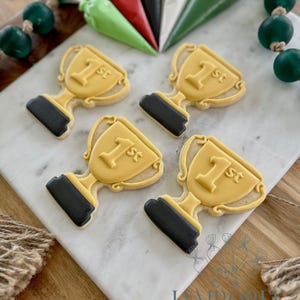 May include: Four trophy-shaped cookies with yellow icing and black bases, each bearing a "1st" inscription. The cookies rest on a white marble surface, accompanied by green, red, black, and white icing bags, and green bead garland.