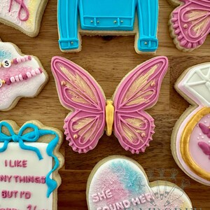 May include: Assortment of decorated sugar cookies with a wedding theme. Includes a blue sweater cookie with "Bride" written on it, a pink butterfly cookie with gold accents, and a heart-shaped cookie with the text "She found her love".