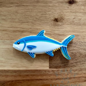 Puede incluir: Una galleta con forma de pez decorada con glaseado azul, blanco y amarillo. La galleta tiene aletas y cola detalladas, sobre un fondo de madera. La galleta es un producto horneado.