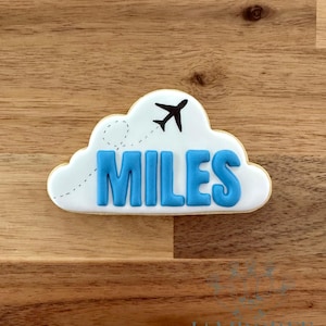 Puede incluir: Una galleta con forma de nube con el nombre "MILES" en letras azules. Un avión negro vuela sobre el nombre, con una línea de puntos que indica su trayectoria. La galleta está sobre una superficie de madera.