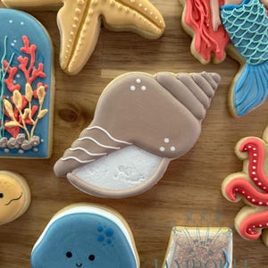 Op de afbeelding: Assortiment van versierde suikerkoekjes met een zeethema. Bevat een zeester, een zeeschelp, een zeemeermin, een kwal en andere oceaan-thema vormen. De koekjes zijn versierd met verschillende kleuren en ontwerpen, waaronder blauw, bruin en oranje.