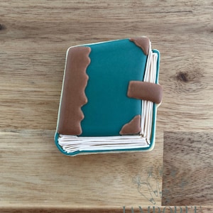 May include: A teal-colored book-shaped cookie with brown accents. The cookie has a detailed design, including a brown clasp and corner pieces. The pages are white, and the edges are outlined in white. The cookie is on a wooden surface.