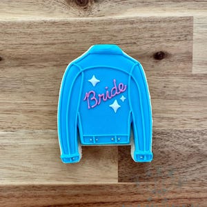 May include: A light blue jacket-shaped cookie with the word "Bride" in pink script on the back. The cookie has white star accents and is set against a wooden background. The cookie is a sweet treat.