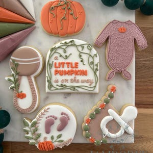 May include: A collection of decorated sugar cookies with a baby shower theme. The cookies include a pumpkin, baby clothes, a rattle, footprints, and a pacifier. The cookies are decorated with orange, green, and pink icing.
