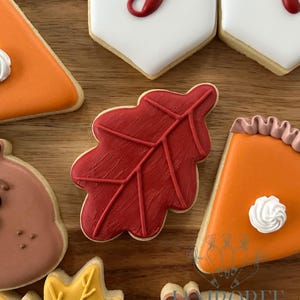 May include: An assortment of decorated sugar cookies in fall shapes. The cookies include a red leaf, pumpkin pie slices, and banner-shaped cookies with red script. The cookies are arranged on a wooden surface.