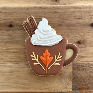May include: A decorated cookie shaped like a brown mug of hot chocolate, topped with white frosting and two cinnamon sticks. The mug features an orange leaf and yellow accents. The cookie is on a wooden surface.