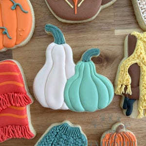 May include: Assortment of autumn-themed cookies, including a white and a teal pumpkin, a fall leaf, a sweater, and a mug. The cookies are decorated with colorful icing and sit on a wooden surface. The cookies are from Home Bakery.