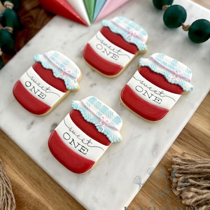 Puede incluir: Cuatro galletas con forma de tarro de mermelada, decoradas con glaseado rojo, blanco y azul. Cada galleta tiene la inscripción "Sweet One". Están dispuestas sobre una superficie de mármol blanco. Elementos decorativos en el fondo.