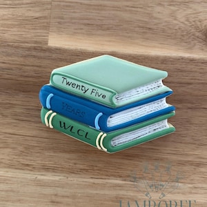 以下が含まれることがあります： 3冊の本の形をしたクッキーの山。一番上の本は薄緑色で「Twenty Five」と書かれています。真ん中の本は青色で「YEARS」と書かれています。一番下の本は緑色で「WLCL」と書かれています。クッキーは木製の表面にあります。