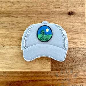 May include: A light blue baseball cap cookie with white stitching and a circular patch featuring a forest scene with a yellow moon. The cookie is on a wooden surface.