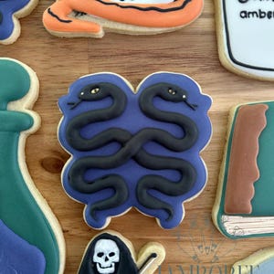 May include: Assortment of Halloween-themed sugar cookies. The cookies feature designs such as a skull, a pair of intertwined snakes, a vial, a book, and a grim reaper playing a violin. The cookies are decorated with black, orange, blue, and white icing.