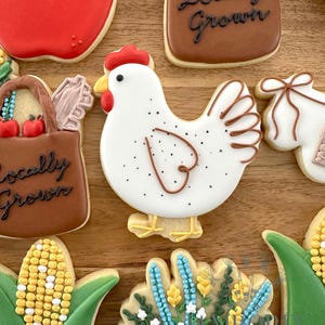 May include: Assortment of decorated sugar cookies on a wooden surface. The cookies include a red apple, a white chicken with brown details, a corn on the cob, and a brown bag with the text "Locally Grown".