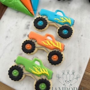 May include: Three colorful monster truck cookies, decorated with blue, orange, and green frosting, feature black tires and yellow flame details. The cookies are arranged on a marble surface, with piping bags in the background.