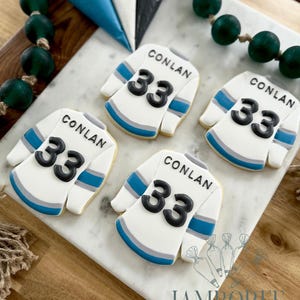 May include: Four white jersey-shaped cookies with black numbers 33 and the name CONLAN. The jerseys have blue and gray accents. The cookies are on a white marble surface, with green beads and piping bags in the background.
