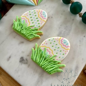 May include: Two Easter egg-shaped cookies with pastel designs and green frosting grass. The cookies are decorated with pink, yellow, and blue patterns. The cookies are on a marble surface.