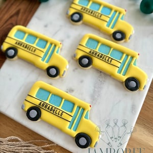 Può includere: Biscotti gialli a forma di scuolabus con finestre blu e il nome "ANNABELLE" in lettere nere. I biscotti hanno ruote nere e fanali posteriori rossi. Sono disposti su una superficie di marmo bianco.