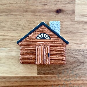 Pode incluir: Uma bolacha em forma de cabana decorada com glacê castanho para imitar paredes de troncos, um telhado azul escuro, uma chaminé azul clara com textura de pedra, uma porta central com puxador preto e uma janela semicircular com detalhes brancos.