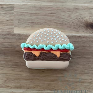 Puede incluir: Una galleta de azúcar decorada con forma de hamburguesa. La galleta presenta un pan de color beige claro con chispas blancas, un volante de lechuga turquesa, tomate rojo, queso naranja y una hamburguesa marrón. La galleta está sobre una superficie de madera.