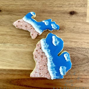 May include: Two sugar cookies shaped like the state of Michigan. The lower cookie is a larger version of the state, and the upper cookie is the Upper Peninsula. Both cookies have a light brown base with blue and white icing.
