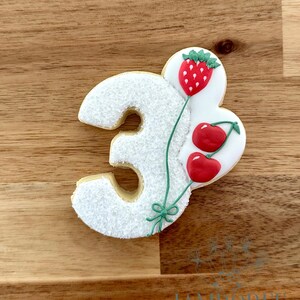 Könnte beinhalten: Ein Keks in Form der Zahl 3, verziert mit weißem Zuckerguss und einem Erdbeer- und Kirschdesign. Die Erdbeere und Kirschen sind rot mit grünen Stielen. Der Keks liegt auf einer Holzoberfläche.