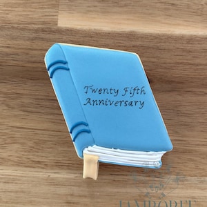 May include: A light blue cookie shaped like a book with the words "Twenty Fifth Anniversary" written on the cover. The cookie has a white edge and a tan bookmark. The cookie is on a wooden surface.