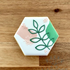 May include: Hexagon-shaped cookie with white icing, featuring a green leaf design and abstract pink and green shapes. Small pink dots and a lace-like design add detail. The cookie sits on a wooden surface.