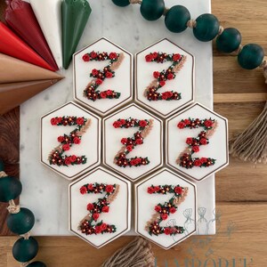 May include: Six hexagonal sugar biscuits with white icing, each decorated with a letter Z formed from piped red roses, green leaves, and tan wheat stalks. The biscuits are arranged on a white marble board with piping bags and dark green beads.