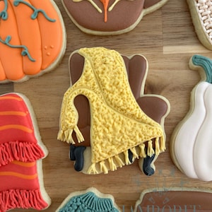 May include: Assortment of decorated sugar cookies with a fall theme. Cookies include a pumpkin, a coffee mug with a leaf design, a chair with a yellow blanket, and a sweater. The cookies are on a wooden surface.