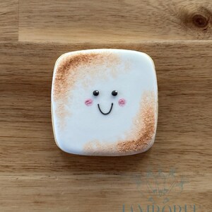 May include: A square cookie designed to look like a toasted marshmallow. It features a white base, a brown toasted edge, black eyes, a curved black smile, and pink cheeks. The cookie rests on a wooden surface.