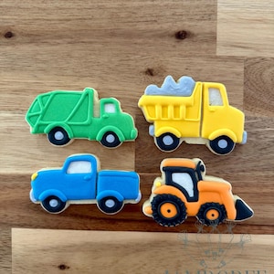 May include: Four vehicle-shaped cookies: a green garbage truck, a yellow dump truck with gray cargo, a blue pickup truck, and an orange tractor with a black blade. Each cookie has black wheels and white accents.