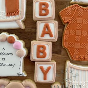 Puede incluir: Surtido de galletas con temática de bebé, que incluyen bloques que deletrean "BABY", una cuna, un body y un oso de peluche. Las galletas están decoradas con glaseado naranja, rosa y blanco. Las galletas están dispuestas sobre una superficie de madera.