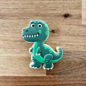 May include: A dinosaur-shaped cookie with green icing and white accents. The cookie features a detailed design with a smiling face, small arms, and a tail. The cookie is decorated with white icing teeth and white icing around the edges.