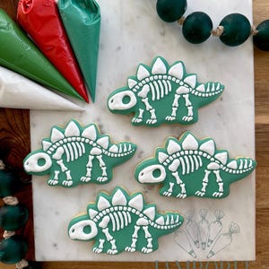 Peut inclure: Quatre biscuits verts en forme de dinosaure avec des détails de squelette blancs sont disposés sur une surface en marbre. Des poches à douille remplies de glaçage rouge, vert et blanc sont visibles. Un brin de perles vertes et le texte "LAMBORELI HOME BAKERY" sont également dans l'image.