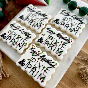 Op de afbeelding: Zes wit geglazuurde koekjes met zwarte letters met de tekst "Happy Birthday & Pixie Prim". De koekjes zijn versierd met groene en rode accenten. De koekjes zijn gerangschikt op een wit marmeren oppervlak.