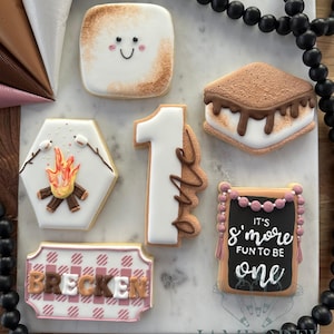 Pode incluir: Uma coleção de biscoitos de açúcar decorados com tema de acampamento. Os biscoitos incluem um marshmallow sorridente, um s'more, uma fogueira, um número um, uma placa com nome e um letreiro que diz "It's s'more fun to be one."
