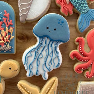May include: An assortment of decorated sugar cookies with a sea life theme. The cookies include a blue jellyfish, a mermaid, a starfish, and an octopus. The cookies are decorated with royal icing in various colors.