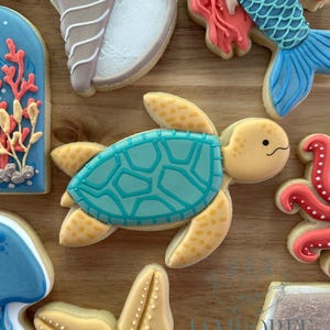 May include: A collection of decorated sugar cookies with a sea life theme. The cookies include a sea turtle with a turquoise shell, a starfish, a mermaid, and other ocean-themed shapes. The cookies are decorated with various colors.