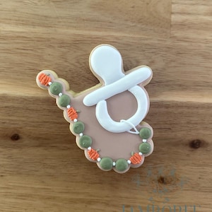 Op de afbeelding: Een baby-thema koekje in de vorm van een fopspeen met een kralenontwerp. De koekje heeft witte glazuur op de fopspeen, een lichtroze basis en groene en oranje kralen details. De koekje ligt op een houten oppervlak.