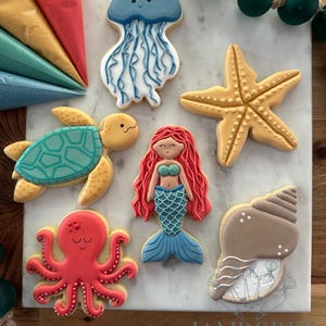 May include: Assortment of ocean-themed cookies, including a jellyfish, starfish, turtle, mermaid, octopus, and seashell. The cookies are decorated with blue, yellow, red, and white icing. The cookies are arranged on a marble surface.