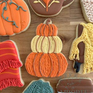 May include: A collection of decorated fall-themed sugar cookies. The cookies include a pumpkin, a coffee mug with a leaf, a sweater, and a scarf. The pumpkin cookie is stacked with orange and yellow icing.