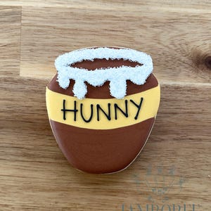 May include: A honey pot cookie with brown and yellow icing, and the word "HUNNY" in black. The top of the cookie has white icing dripping down. The cookie is on a wooden surface.