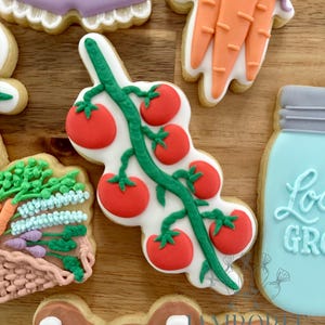 May include: Assortment of decorated sugar cookies, including a cluster of red tomatoes on a green vine, a carrot, a basket of vegetables, a jar, and a cookie with the word "baby". The cookies are on a wooden surface.