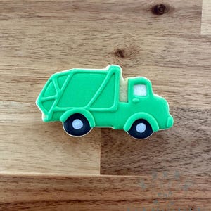 Może przedstawiać: Zielone ciastko w kształcie śmieciarki z czarno-białymi kołami. Ciastko ma biały kontur i jest umieszczone na drewnianym tle. Ciężarówka ma szczegółowy projekt nadwozia.