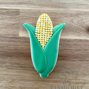 Puede incluir: Una galleta con forma de mazorca de maíz con un diseño de granos amarillos y blancos, envuelta en hojas de color verde brillante. La galleta tiene un contorno blanco y está sobre un fondo de madera. El texto "JAMBOREE Home Bakery" es visible.