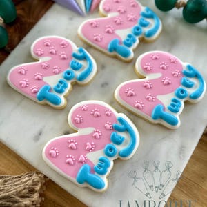 May include: Four heart-shaped cookies decorated with pink icing, paw prints, and the word "Two" in blue icing. The cookies are on a white marble surface, with piping bags and decorative beads in the background. The cookies are from Jamboree Home Bakery.