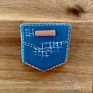 May include: A blue cookie decorated to resemble a denim pocket. The cookie features white stitching details, a small pink patch, and a white border. The cookie is set against a wooden background.